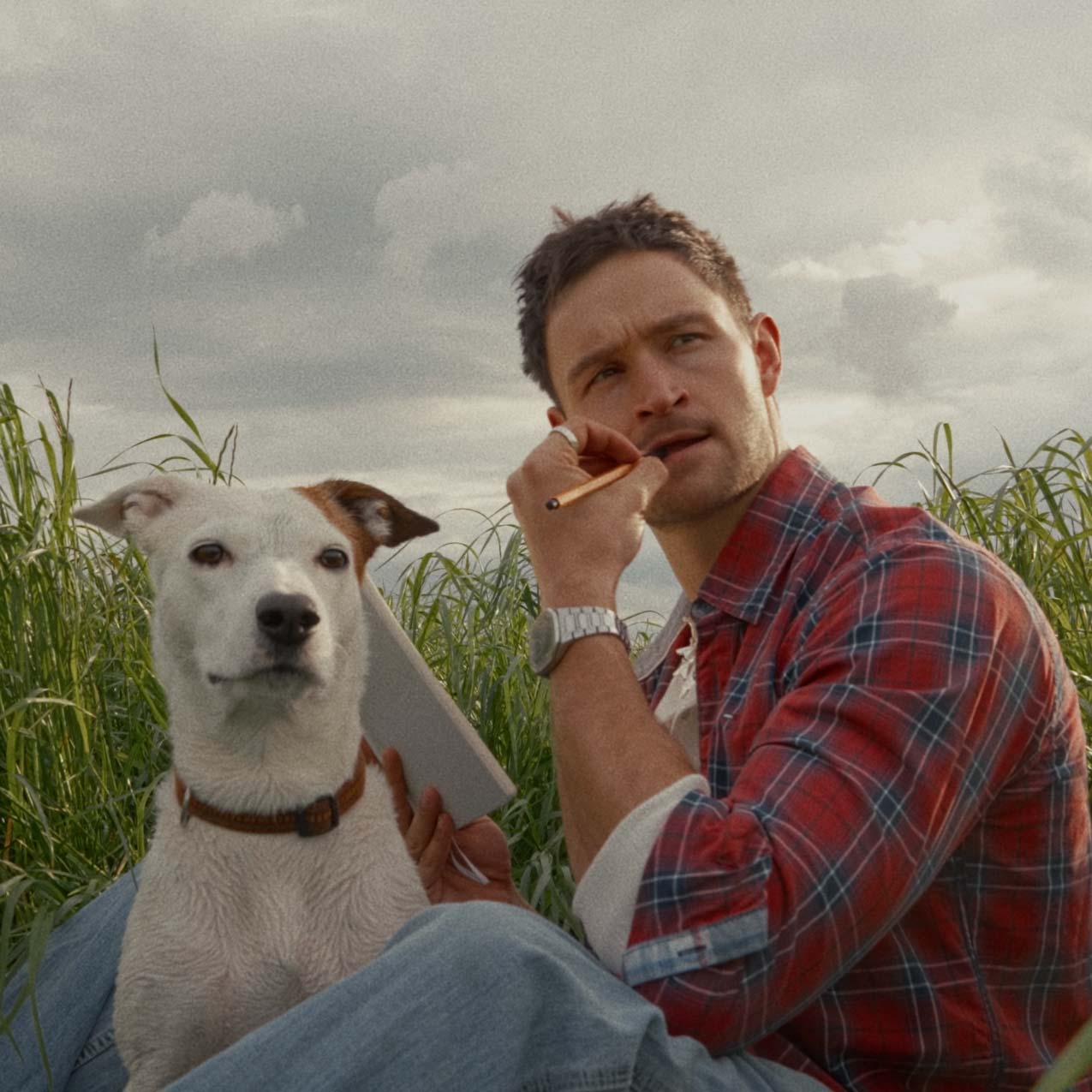 Nick sitting in a field with his dog Zala, pen in hand, thinking